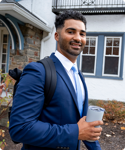man wearing backpack and holding a coffee cup walking outside and smiling at the camera