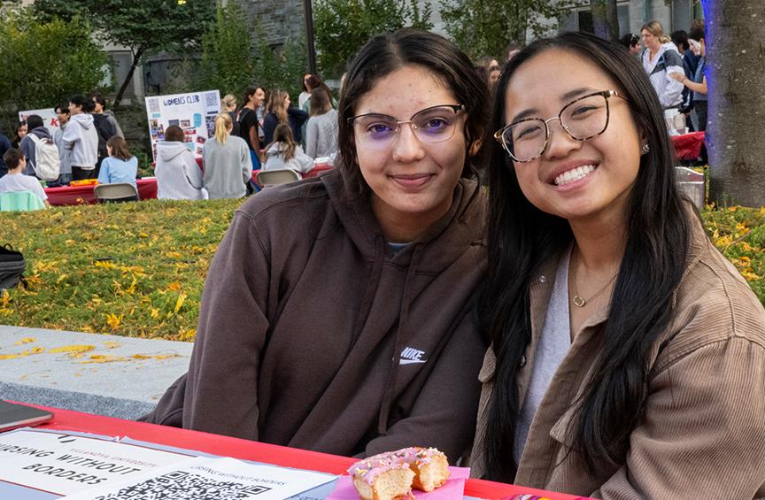 Two female friends smile directly at the camera sitting at a table outside. 