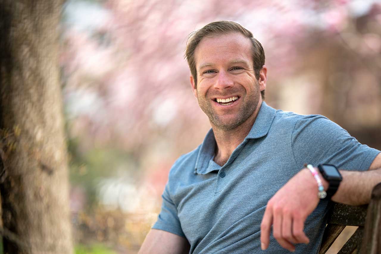 Sean Wade smiles while sitting on a bench outside, with a blurred background of pink blossoms on a tree.
